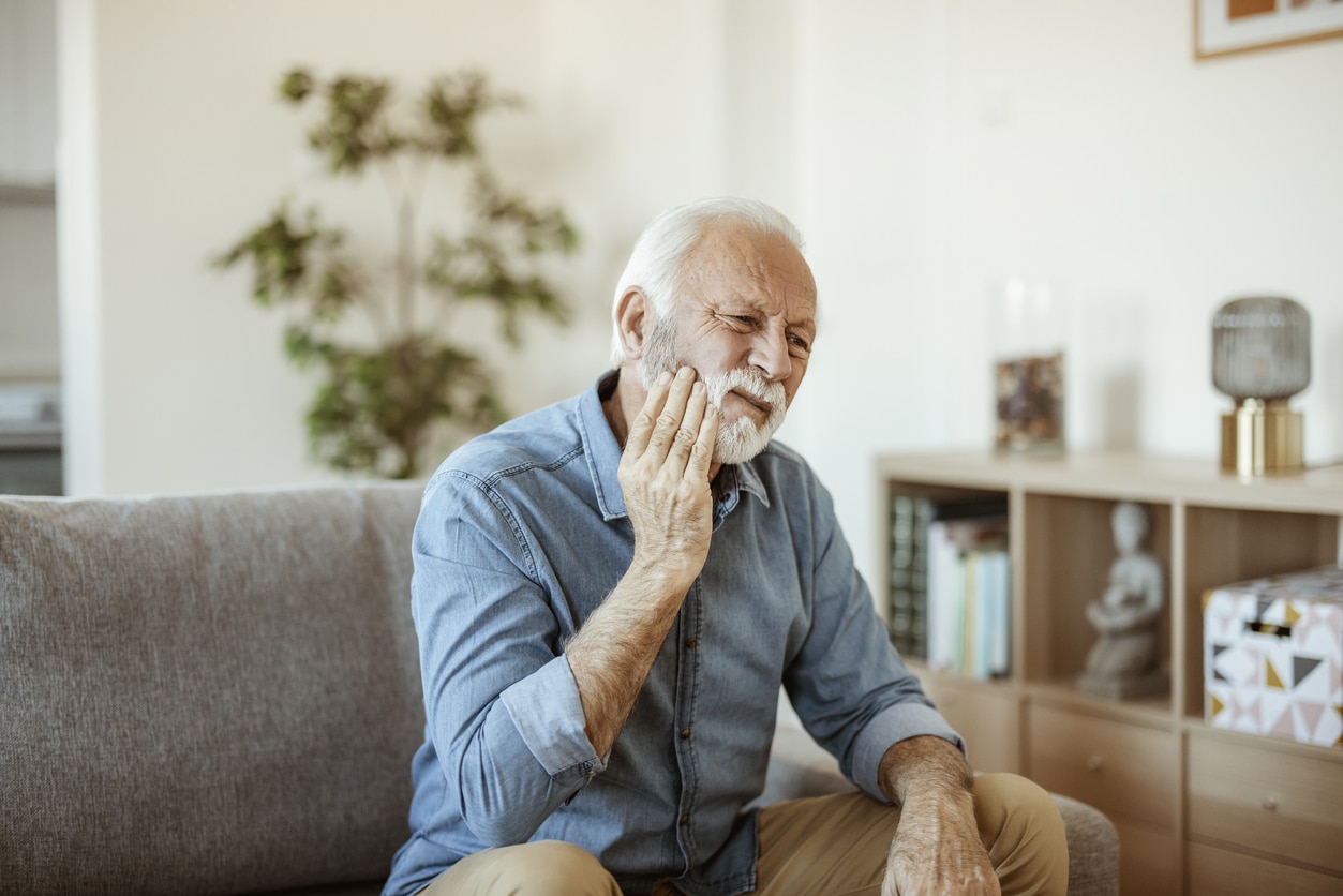 A man rubs his jaw. The man is sitting on a couch, is wearing a blue denim shirt, and has a white beard.